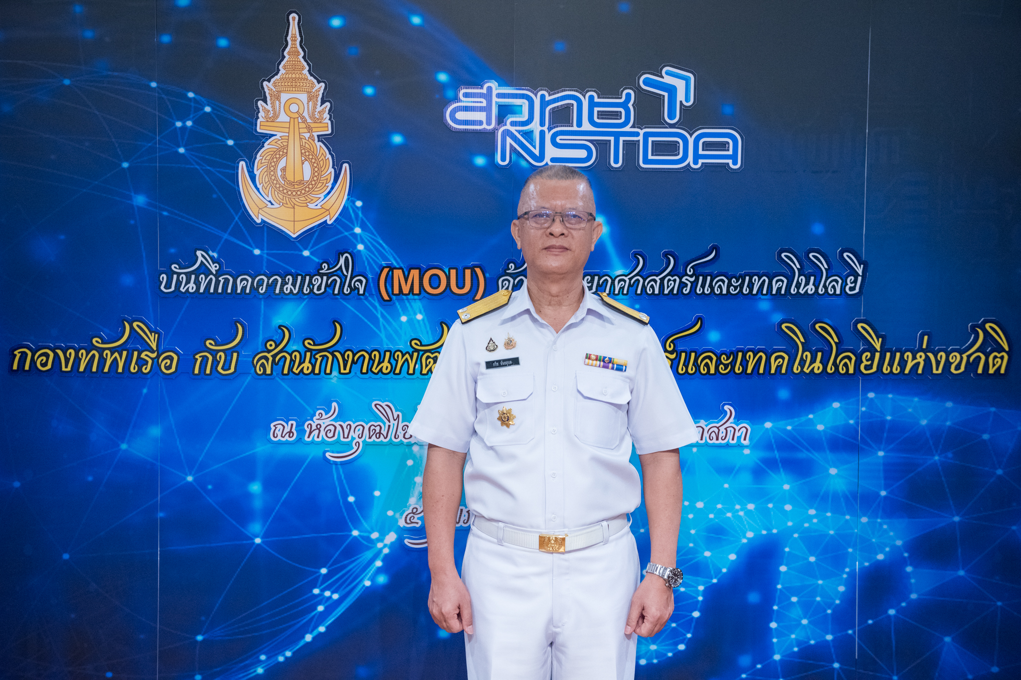 พลเรือตรี กริช ขันธอุบล เจ้ากรมวิทยาศาสตร์ทหารเรือ ยืนบนเวทีที่แสดงข้อความบันทึกความร่วมมือด้านการวิจัยและพัฒนาทางวิทยาศาสตร์และเทคโนโลยี (MOU)